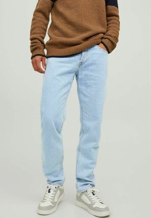 Hombre usando un suéter de punto marrón, jeans azul claro y zapatillas blancas y grises, con una mano en el bolsillo, de pie contra un fondo sencillo.