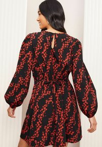 Robe noire avec un motif floral rouge, manches bouffantes longues, taille élastique et détail à fermeture en forme de clé au dos. Confectionnée en tissu léger.