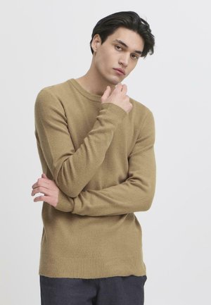Suéter marrón claro con mangas largas, cuello redondo, puños y dobladillo acanalados, hecho de un material suave, llevado por un modelo frente a un fondo neutro.