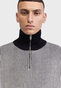 Pulover tricotat cu guler înalt dintr-un material rib elastic negru, închidere cu fermoar și un model vertical cu dungi alb-negru. Textură netedă.