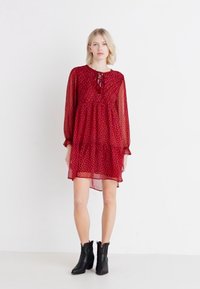 Rochie roșie, transparentă, cu mâneci lungi și o croială lejeră, cu volan la tiv și detaliu cu fundă la gât. Puncte mici albe în toată rochia.