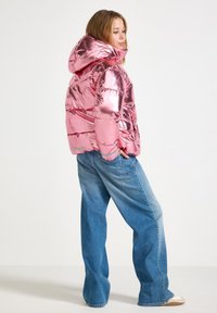 Woman standing sideways wearing a shiny pink puffer jacket with hood, loose blue jeans, and white sneakers against a plain background.