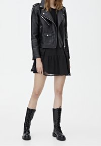 Femme portant une veste de motard en cuir noir, une robe courte noire et des bottes noires à lacets montant jusqu'aux genoux, debout sur un fond blanc.