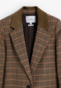 Blazer de cuadros marrón con cuello de ante marrón y botones negros. Presenta un diseño clásico de solapa y un forro interior oscuro. Hecho en España.