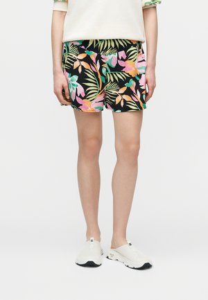 Személy fekete rövidnadrágot visel színes trópusi levélmintával, világos bézs felsővel és fehér, fekete talpú slip-on cipővel.