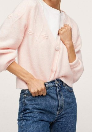 Kvinde iført lyserød cardigan med knapper over hvid top, justerer højtaljede blå jeans med hånden i lommen.