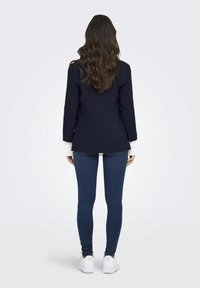 Granatowa marynarka z długimi rękawami, zestawiona z białą koszulą, ciemnymi dżinsami i białymi trampkami. Gładki materiał z dopasowanym krojem.