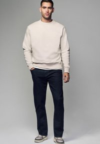 Sudadera de color beige claro con puños y dobladillo acanalados, combinada con jeans azul oscuro y zapatillas grises. Diseño sencillo, textura de tela resistente.