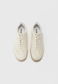 Hvite semskede sneakers med beige gummisåler, med en glatt tekstur, rund tå og klassisk snøresystem. Merkenavn synlig inni.
