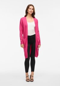 Lange roze cardigan van zachte stof met ribgebreide manchetten en twee voorkruiszakken, gecombineerd met een witte top en zwarte broek.