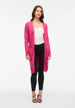 Lang rosa cardigan laget av myk stoff med ribbede mansjetter og to frontlommer, kombinert med en hvit topp og svarte bukser.