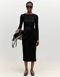 Zwarte jurk met lange mouwen en een geknoopte taille. Model houdt een donker bordeauxrood tasje vast. Glad materiaal met een aansluitende pasvorm. Zwarte schoenen.