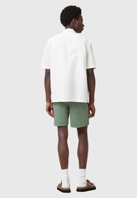 Camicia bianca a maniche corte, vestibilità rilassata, abbinata a pantaloni corti verdi oliva. I dettagli includono una spalla posteriore e una leggera fessura laterale.