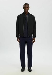 Giacca bomber nera con zip, polsini a costine e una camicia scura sotto, abbinata a pantaloni blu navy e scarpe nere.