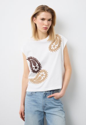 Giovane donna che indossa una canotta bianca con motivi paisley marroni e beige e jeans azzurri, in piedi contro un muro bianco.