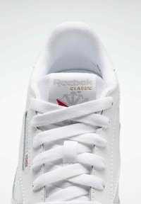 Fehér Reebok sportcipő textil és bőr anyagból, kerek orral, fehér fűzővel, logóval a nyelvén és piros akcentussal a saroknál.