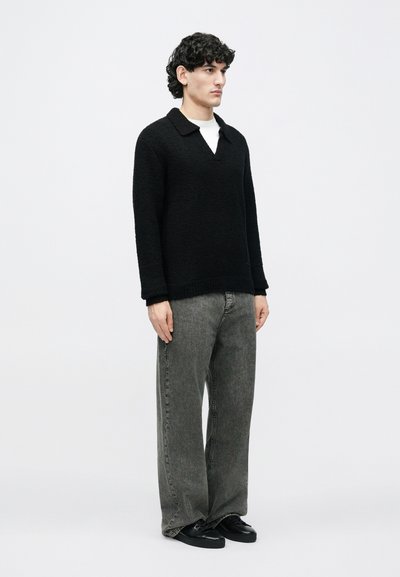Bărbat în picioare, purtând un pulover texturat negru cu mâneci lungi, blugi gri largi, pantofi negri și un tricou alb pe sub pulover, pe un fundal simplu.