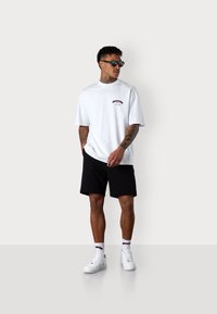 T-shirt en coton blanc à manches courtes avec un petit logo rouge, associé à un short noir, des baskets blanches et des lunettes de soleil noires.