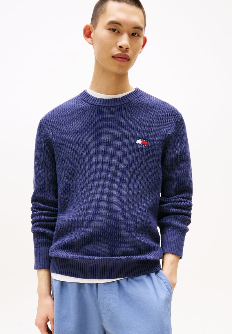 Maglione a coste navy con colletto rotondo e piccolo logo. Maniche lunghe con polsini a costine e vestibilità rilassata. Indossato sopra una camicia bianca.