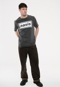 Gray cotton t-shirt with a prominent white "oasis" logo, paired with dark brown loose-fitting pants and black shoes. Clear, simple design.