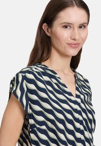 Bluzka z krótkim rękawem w kolorze granatowym z falistym wzorem w białym i zielonym kolorze. Projekt w serek, luźny krój oraz miękka, gładka faktura.