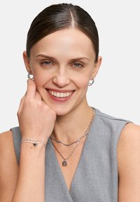 Sølv smykker herunder hoops øreringe, en bølget choker og lagdelte halskæder med charms på en person iført en grå top.