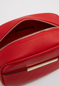 Punainen nahkalaukku, jossa tikattu kuvio, hopeavetoketju ja kultaiset metalliosat. Avoin yläosa paljastaa pehmeän sisuksen, jossa on merkki.