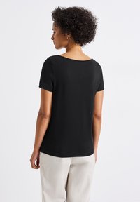 Mustapaitainen lyhythihainen paita, jossa on pyöreä pääntie ja joka on valmistettu sileästä materiaalista. Selkäosa on yksinkertainen ja rento malli, yhdistettynä vaaleanbeigeihin housuihin.