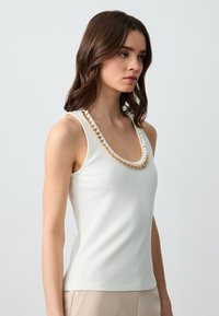 Top a canotta bianco a coste con scollatura profonda a barca, caratterizzato da dettagli ornamentali in catene dorate lungo il collo. Texture liscia, design aderente.