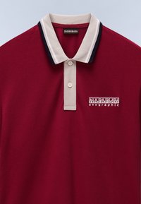 Polo bordeaux con colletto a righe crema e navy, pattina con tre bottoni e logo ricamato "Napapijri geographic". Tessuto strutturato.