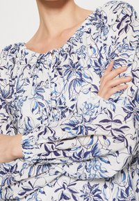 Blouse florale avec un fond blanc et des motifs floraux bleus et marine, encolure élastiquée et poignets avec une texture froncée.