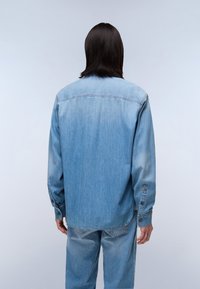 Lyseblå denimskjorte med lange ærmer, knappede manchetter og en afslappet pasform. Har en lige kant og subtil falming for tekstur.