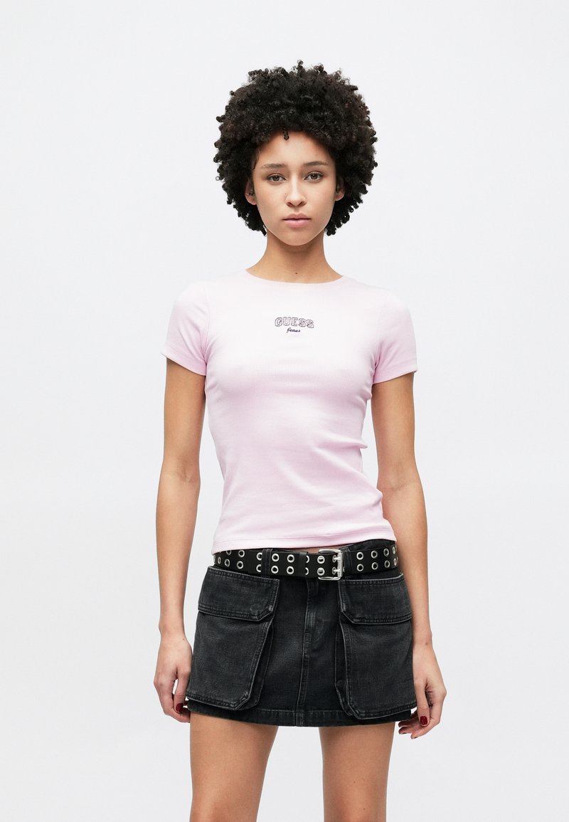 T-shirt rosa ajustada com mangas curtas e logótipo "GUESS", combinada com uma saia de ganga preta com bolsos e um cinto com tachas e ferragens prateadas.