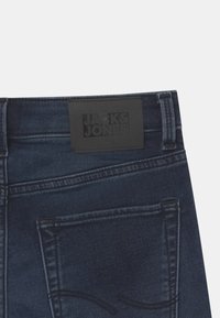 Jeans de denim azul oscuro con un parche negro etiquetado "JACK & JONES" en la cinturilla, que presentan bolsillos estándar y costuras contrastantes.