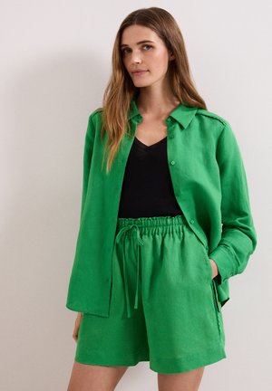 Vrouw met lang bruin haar draagt een felgroen overhemd met knopen en bijpassende shorts met een zwart topje eronder, staand tegen een vlakke muur.