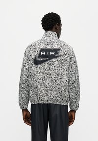 Μπουφάν Nike σε ασπρόμαυρο με στίγματα, με ψηλό γιακά και μεγάλο μαύρο λογότυπο "AIR" στην πλάτη. Ελαφρύ υλικό με ελαστικές μανσέτες.