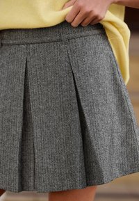 Pelēka houndstooth raksta plīsēta svārki ar teksturētu audumu. Tam ir jostasvietas lenta un tie sniedzas virs ceļa, kombinēti ar dzeltenu topu.