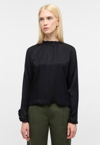 Blusa nera in satin a maniche lunghe con colletto e polsini arricciati; presenta una vestibilità comoda e una superficie liscia. Indossata con pantaloni verde oliva.