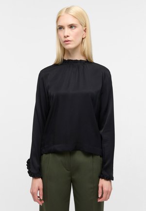 Schwarze Satinbluse mit langen Ärmeln, gerafftem Ausschnitt und Manschetten; hat eine lockere Passform und eine glatte Textur. Kombiniert mit olivgrünen Hosen.