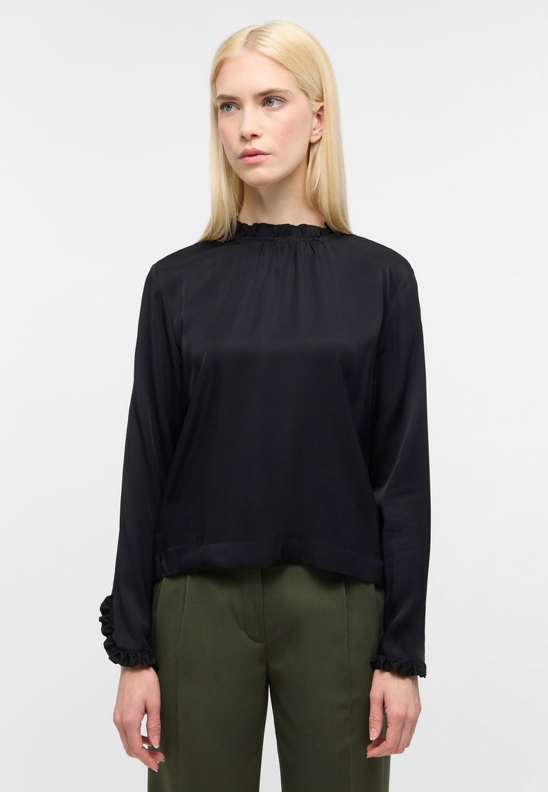Schwarze Satinbluse mit langen Ärmeln, gerafftem Ausschnitt und Manschetten; hat eine lockere Passform und eine glatte Textur. Kombiniert mit olivgrünen Hosen.
