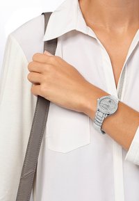 Sølvur med en rund urskive og funklende detaljer på en metalrem, båret på et håndled med en lys beige bluse og en grå taske-rem.