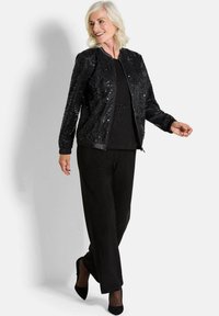 Veste bomber noire avec des embellissements en sequins, des poignets côtelés et une fermeture éclair, associée à un pantalon noir ample et des talons.