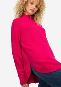 Maglione zip-up fucsia con colletto alto, fessure laterali e polsini a coste. Texture morbida, vestibilità ampia e maniche lunghe. Abbinato a un paio di jeans.