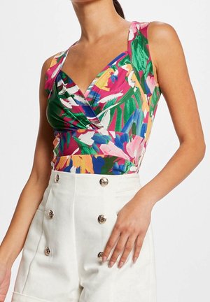 Frau trägt ein buntes, ärmelloses Blumenoberteil, das in eine hoch taillierte weiße Hose mit sechs dekorativen goldenen Knöpfen gesteckt ist.