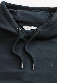 Sudadera con capucha en tela de un azul marino oscuro, con un cordón con puntas metálicas y un pequeño logo de un ciervo en la parte delantera izquierda.
