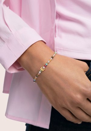 Pulsera de cuentas con piedras multicolores, detalles dorados y una letra "E" en dorado. Se lleva en la muñeca, con una camisa de color rosa claro visible.