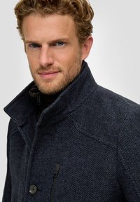 Marineblauwe wolmix jas met een hoge kraag, knoopsluiting en een textuurstof met een subtiel patroon. Zijzak met ritsdetail.