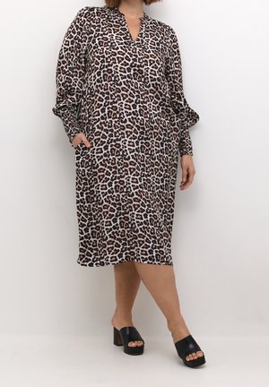Woman wearing knee-length leopard print dress with long sleeves and black open-toe block heel sandals, standing with one hand in pocket.