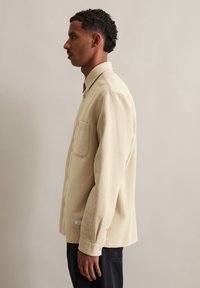 Beige blouse met lange mouwen in een gestructureerde stof, voorzien van een borstzak en knoopsluiting. Heeft een kraag en aansluitende manchetten.