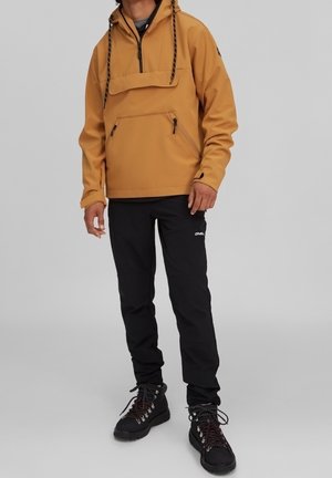 Mężczyzna w musztardowożółtej kurtce z kapturem i kieszeniami na zamek, czarnych spodniach i czarnych butach turystycznych na szarym tle.